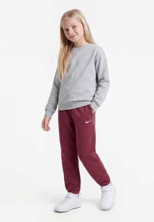 Jeune fille aux longs cheveux blonds debout, portant un sweat-shirt gris, un pantalon de survêtement Nike bordeaux et des baskets blanches sur fond blanc.