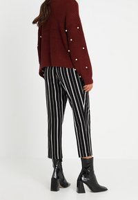 Burgundská pletená svetr s perlovými akcenty, spárovaný s černobíle pruhovanými zkrácenými kalhotami a černými kotníkovými botami s masivním podpatkem.