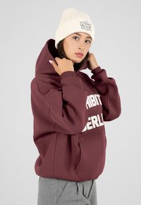 Joven mujer ajustando una sudadera marsala con texto blanco, llevando un gorro de punto crema y pantalones grises, de pie frente a un fondo claro y liso.