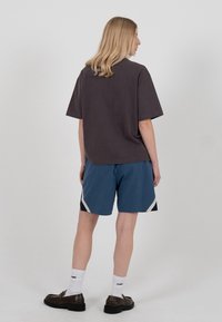 Personne blonde vue de dos, portant un t-shirt ample foncé, un short bleu avec des accents blancs, des chaussettes blanches montantes et des mocassins marron.