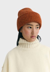 Junge Person mit gerippter, burnt orangefarbenen Mütze und cremefarbigem, geripptem Rollkragenpullover, die zur Seite schaut, vor einem schlichten Hintergrund.