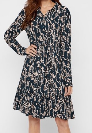 Kobieta ubrana w długą sukienkę z kołnierzykiem i rozpinaną na guziki, wykonana z abstrakcyjnej tkaniny w czarno-beżowy wzór, z ręką na biodrze, stojąca na białym tle.