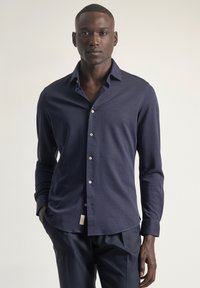 Camisa de manga larga en tela lisa color azul marino, con cierre de botones en la parte frontal, cuello puntiagudo y detalle sutil de logo. Corte entallado y dobladillo redondeado.