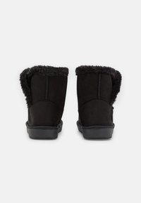 Botas de tobillo negras con un forro de piel de peluche alrededor de la parte superior, detalles de costura visibles y suelas de goma planas, mostradas desde la parte de atrás sobre un fondo blanco.