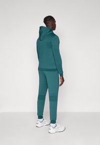 Teal hettegenser og matchende joggebukser, med ribbestrikkede mansjetter og knepplaster. Hvite sportssko med blå detaljer fullfører antrekket.