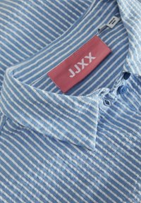 Απλί μπλε πουκάμισο με λεπτές λευκές ρίγες, βαμβακερό ύφασμα, γιακά με κουμπιά και ροζ ετικέτα με το όνομα της μάρκας "JJXXX".