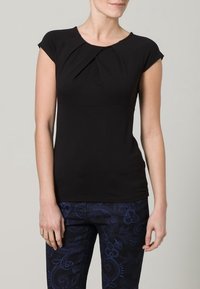 Svart kortärmad topp med veckad halsringning, fitted silhuett och slät textur, ihopparad med mörka byxor med ett blått paisleymönster.