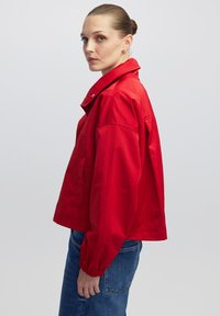 Vrouw met slicked-back haar die gouden oorringen draagt, een rode colbert en een spijkerbroek, staande tegen een effen lichte achtergrond.