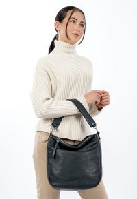 Musta nahkainen olkalaukku, jossa on kaareva yläosa ja säädettävä hihna, vetoketjullinen suljin ja minimalistinen muotoilu. Sileä pinta.