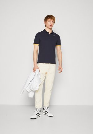 Polo navy con dettagli gialli, indossato con pantaloni crema e sneaker in bianco e nero, mentre tiene una giacca bianca. Design semplice, vestibilità classica.