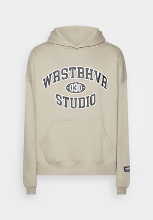 Beige hoodie met voorvak en zwarte tekst "WRSTBHVR 030 STUDIO" op de borst, geribbelde boorden en klein zwart label op de mouw.