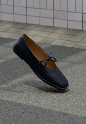 Scarpa piatta in pelle nera con punta quadrata e fiocco annodato, sospesa sopra un marciapiede grigio con sfondo in piastrelle.