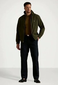 Homme portant une veste vert foncé sur une chemise marron, un pantalon noir et des chaussures noires, se tenant avec une main dans la poche en regardant sur le côté.