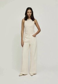 Beige sleeveless top paired with wide-leg cream pants. Smooth fabric, fitted bodice, high waist, and subtle pocket detail. Pointed beige shoes.