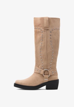 Beżowy zamszowy kozak do połowy łydki z srebrnymi ćwiekami, ozdobnym paskiem i kółkiem wokół kostki oraz niskim czarnym obcasem typu słupek.