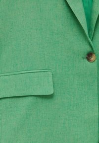 Groene linnen blazer met een gekleed kraag, enkele knoopsluiting en een klepzak. De stof heeft een textureel, geweven uiterlijk.