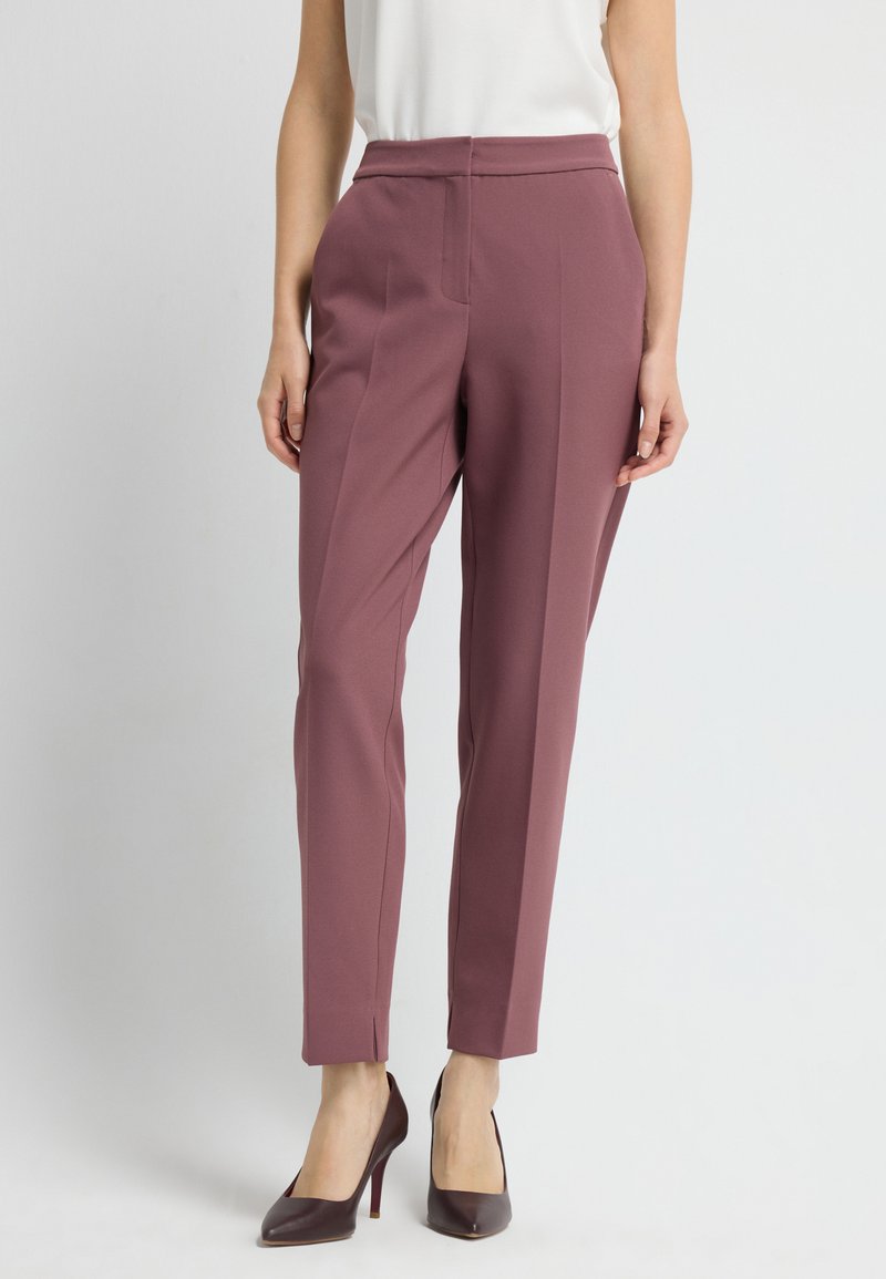 Broek van gedempt paarse stof, getailleerde pasvorm, taps toelopende pijpen, plooien aan de voorkant en zijzakken; gedragen met donkergekleurde hakken.