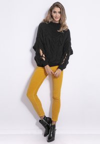 Czarny dzianinowy sweter z wzorem w warkocze i wyciętymi rękawami, zestawiony z musztardowymi spodniami z kurczaka oraz czarnymi botkami za kostkę z ćwiekami.