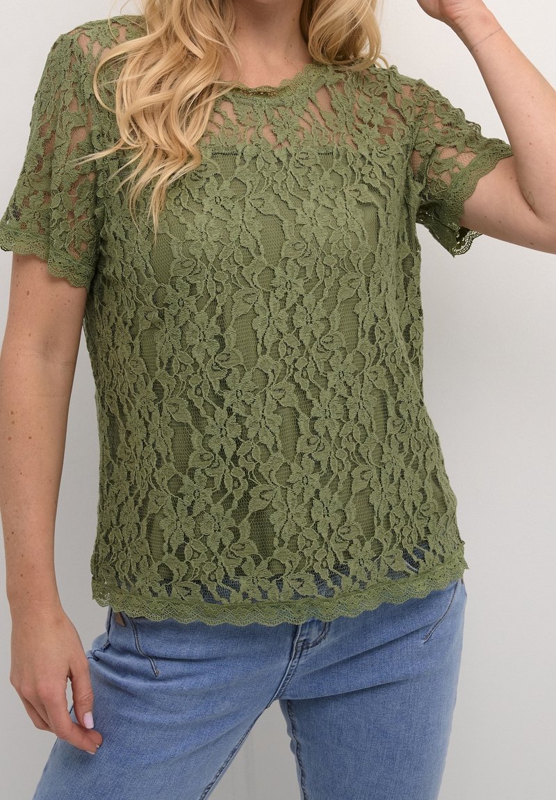 Haut en dentelle vert olive à manches courtes, bords festonnés, motifs floraux et texture semi-transparente, porté avec un jean bleu.