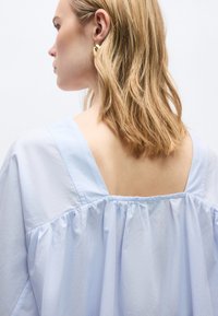 Γυναίκα με ξανθά μαλλιά μήκους μέχρι τους ώμους φοράει ανοιχτό γαλάζιο φαρδύ πουκάμισο με φαρδύ, τετράγωνο ντεκολτέ που αφήνει ακάλυπτη την άνω πλάτη, εμφανίζοντας χρυσό κρίκο σκουλαρίκι.