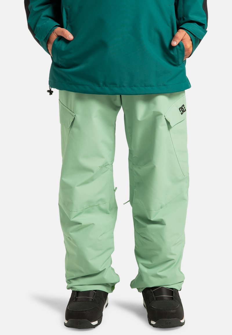 Miętowe spodnie narciarskie o gładkiej teksturze, z bocznymi kieszeniami i logo. Połączone z czarnymi butami narciarskimi.