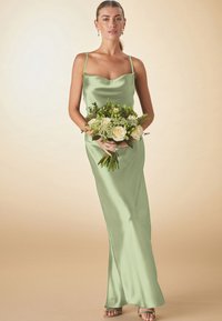 Femme en robe longue en satin vert clair tenant un bouquet de roses blanches et de verdure, debout devant un fond beige uni.