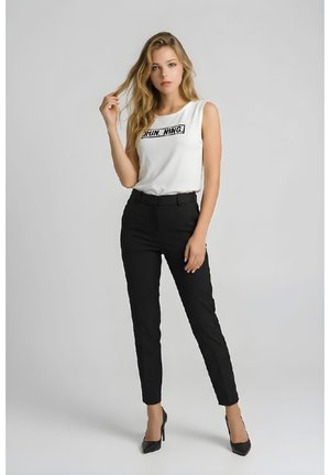 Mujer rubia con una blusa blanca sin mangas con el texto "RUNNING", pantalones negros ajustados y tacones altos negros, de pie contra un fondo gris.