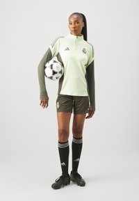 ADIDAS futbola apģērbs: gaiši zaļa garām piedurknēm blūze, tumši zaļas šortus, melnas ceļgalu augstās zeķes ar baltām svītrām, melni futboli un tur rokā futbola bumbu.
