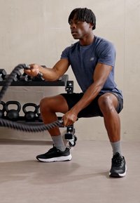 Corde de fitness noire, t-shirt de sport gris, shorts noirs, chaussettes grises et chaussures noires. Équipement de poids visible en arrière-plan.