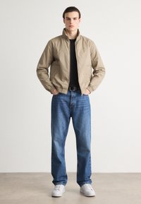 Beige bomber-takki, jossa on vetoketju edessä ja elastiset hihansuut, yhdistettynä sinisiin suoriin farkkuihin ja valkoisiin lenkkareihin. Yksinkertainen muotoilu ja rento istuvuus.