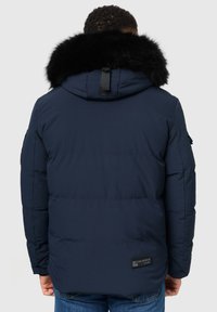 Tamno plavi puffer jakna s odvojivim crnim krznom od umjetne kože, zatvaranje na patentni zatvarač i horizontalnim šavovima. Ima logotip na donjem dijelu leđa.