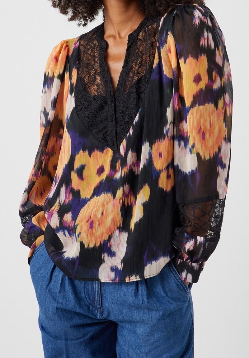 Femme portant un chemisier floral coloré avec un col en V en dentelle noire et des poignets, associé à un pantalon en jean bleu ample.