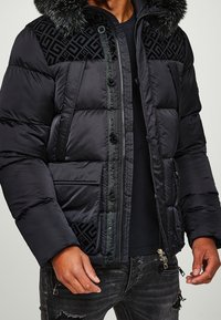 Mand med en sort dunjakke med pelskrave, geometrisk mønster på skuldrene, forlommer og lynlåsede sorte slidte jeans.