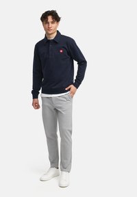 Polo a maniche lunghe blu navy realizzato in tessuto morbido, con un colletto a tre bottoni e un logo rosso. Abbinato a pantaloni tapered grigio chiaro e sneakers bianche.