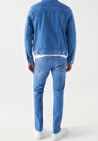 Muž ve světle modré džínové bundě a džínách s bílými teniskami, stojící proti jednoduchému bílému pozadí, zády k fotoaparátu.