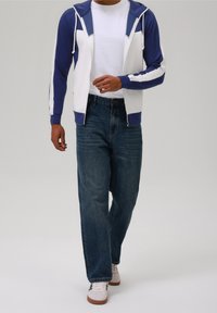 Felpa con cappuccio e zip blu e bianca con coulisse, abbinata a jeans a gamba dritta blu scuro e sneakers bianche. Abbigliamento atletico casual.