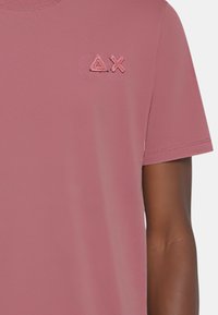 T-shirt rosa in cotone a maniche corte con scollatura rotonda e loghi a triangolo e croce ricamati di un rosa più chiaro sul petto.