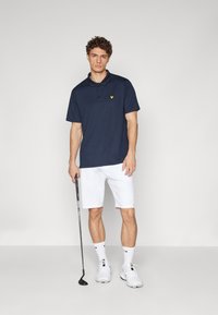 Námořnická modrá polo tričko so žltým logom, kombinované s bielymi šortkami. Model drží golfovú palicu, má na sebe biele tenisky a ponožky.