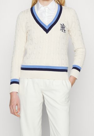 Frau trägt einen cremefarbenen Pullover mit Zopfmuster und V-Ausschnitt, mit blauen und marineblauen Streifen, Monogramm auf der Brust, darunter ein weißes Hemd und weiße Hose.