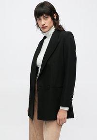 Blazer negro de doble botonadura con solapas de pico, hombros estructurados y bolsillos laterales. Se lleva sobre un jersey de cuello alto claro y se combina con pantalones texturizados.
