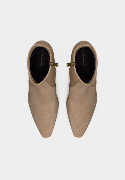 Ζεύγος μπεζ σουέντ μποτάκια αστραγάλου με μυτερά δάχτυλα και κοντά χρυσά φερμουάρ στις εσωτερικές πλευρές, εμφανιζόμενα από πάνω σε λευκό φόντο.