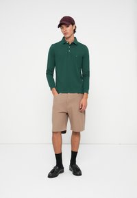 Jongeman in een donkergroen poloshirt met lange mouwen, beige shorts, zwarte sokken, zwarte instappers en een bordeauxrode pet, staand tegen een eenvoudige witte muur.