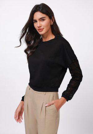 Svart genser med kort, løs passform og blonder på ermene. Kombinert med beige høyhalsede bukser med sidelommer.