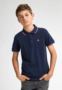 Námořnická modrá polo košile s úzkým střihem, krátkými rukávy, límečkem a červenými pruhovanými akcenty na límci a manžetách; obsahuje malé logo.