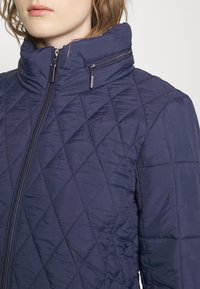 Granatowa pikowana kurtka z wysokim kołnierzem, zamkiem błyskawicznym i teksturowanym wzorem w kształcie diamentu na tkaninie syntetycznej.