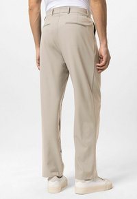 Beige broek met een rechte pijp, voorzien van twee achterzakken en subtiele ritsaccenten. Draagt witte lage sneakers.