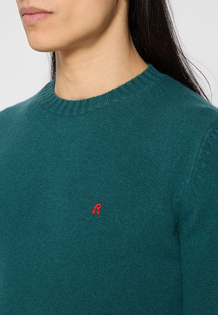Teal gebreide trui met een ronde halslijn en geribbelde manchetten, voorzien van een klein rood "R"-logo op de linker borst. Zachte textuur.