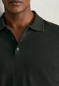 Tmavě zelené polo tričko s límečkem, dvoubarevným zapínáním na knoflíky a měkkou texturou. Detailní záběr zdůrazňující strukturu látky a padnutí.