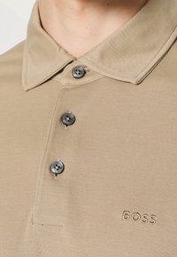 Gros plan d'un polo beige avec trois boutons foncés et un logo "BOSS" brodé sur la poitrine.