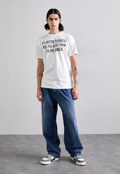 White cotton t-shirt featuring bold black text, paired with loose blue denim jeans and gray sneakers with black and white accents.
